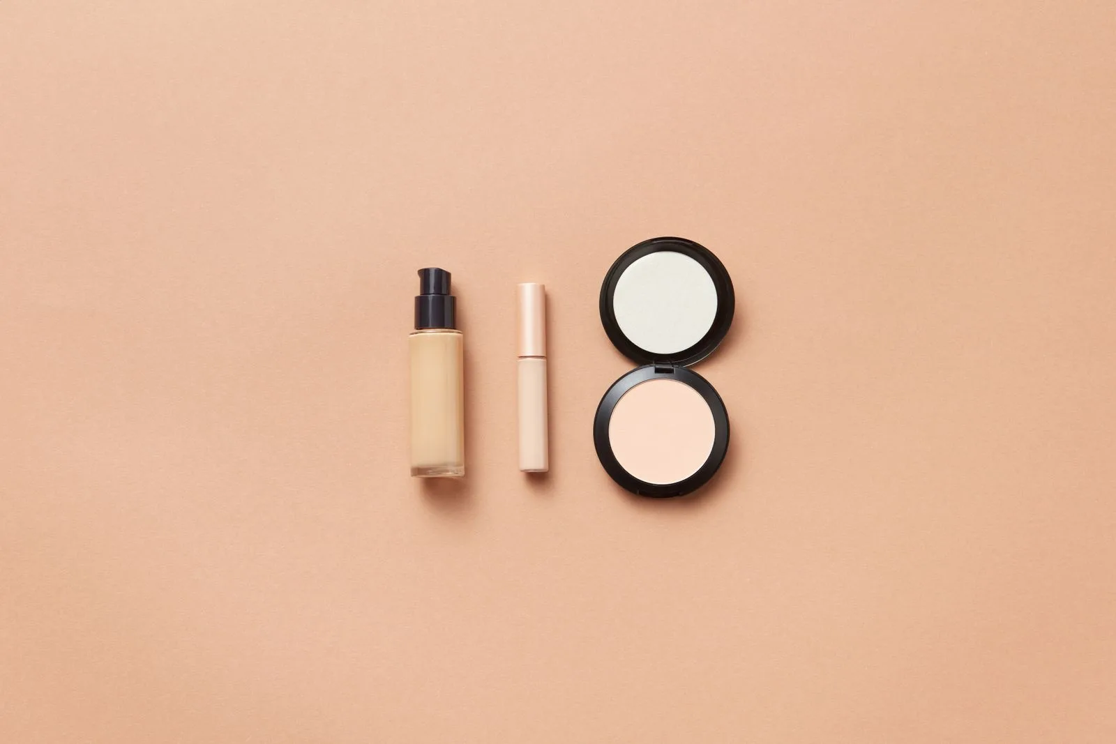 A bottle of liquid foundation, a concealer and an open compact powder on a beige background
